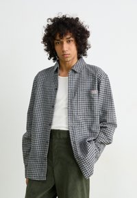 Jongere met kroeshaar die een septumring draagt, een wit tanktop, een geruite blouse en donkergroene broek, staande met hand in pocket.