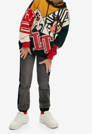 Multikleurige hoodie met cartoons en gedurfde graphics, gecombineerd met grijze broek en witte sneakers. Rode accenten op de manchetten.