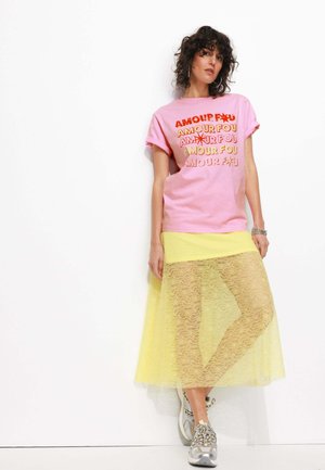 T-shirt en coton rose avec le texte "AMOUR FOU" en orange et jaune. Jupe en dentelle jaune superposée. Baskets grises avec des accents argentés.