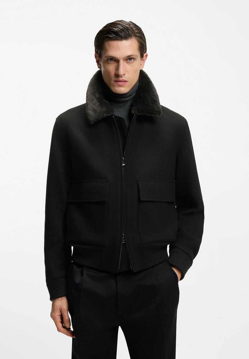 Mann trägt eine schwarze Jacke mit Reißverschluss und großen Fronttaschen sowie einem dunklen Fellkragen, kombiniert mit schwarzen Hosen und einem dunklen Rollkragenpullover.