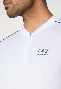 Λευκή αθλητική μπλούζα με φερμουάρ, φτιαγμένη από πλεκτό ύφασμα με πλέγμα, με όρθιο για κά, μπλε περίγραμμα και λογότυπο EA7 Emporio Armani στο αριστερό στήθος.