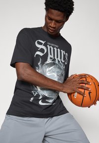 Czarny t-shirt z grafiką "San Antonio Spurs" i wzorem koszykarskim, w połączeniu z jasnoszarymi szortami, trzymający pomarańczową piłkę do koszykówki.