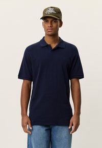 Μπλε ναυτικό πόλο μπλουζάκι με κλασικό γιακά και κοντά μανίκια, κατασκευασμένο από βαμβάκι. Συνδυασμένο με πράσινο ελιάς καπέλο με γραφικό σχέδιο.