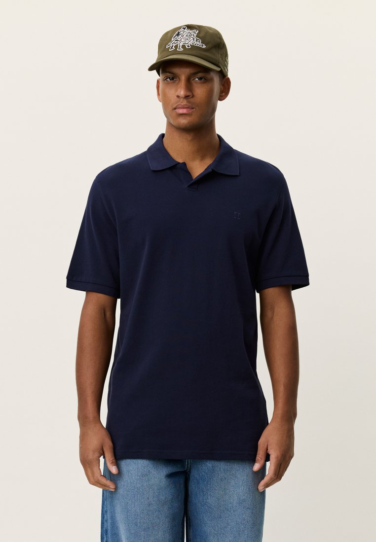 Μπλε ναυτικό πόλο μπλουζάκι με κλασικό γιακά και κοντά μανίκια, κατασκευασμένο από βαμβάκι. Συνδυασμένο με πράσινο ελιάς καπέλο με γραφικό σχέδιο.