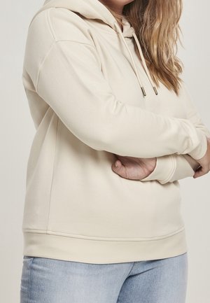 Frau mit hellbraunen Haaren, die einen cremefarbenen Kapuzenpullover und hellblaue Jeans trägt, mit verschränkten Armen vor dem Oberkörper, neutraler Hintergrund.