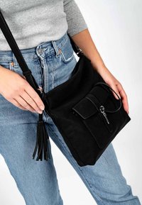 Schwarze Umhängetasche aus Stoff mit strukturierter Oberfläche, Fronttasche und Quasten-Akzent, gehalten von einer Person, die helle Jeans aus Denim trägt.