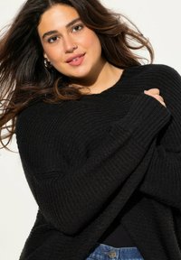 Svart stickad tröja med lös passform och ribbade ärmar. Buren över en svart topp, ihop med blå denim som har ett texturerat mönster.