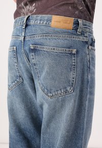 Jean en denim bleu clair avec une coupe décontractée, comprenant deux poches arrière, une texture délavée et une étiquette en cuir beige au niveau de la taille.