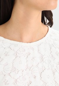 Top en dentelle blanche présentant des motifs floraux, un col rond et une surface texturée. Le tissu est léger et semi-transparent.