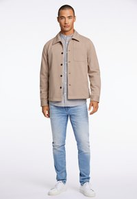 Giacca beige con bottoni e tasca a toppa, abbinata a una camicia a righe e jeans azzurri chiari, completata da sneakers bianche.