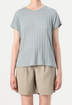 Person iført en lyseblå ribbet kortærmet top og beige knælange shorts, stående mod en ensfarvet hvid baggrund.