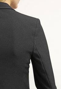 Veste noire sur mesure en tissu lisse, avec épaules structurées et détails de couture soignés. La coupe ajustée met en valeur la forme du dos.