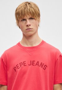 Κόκκινο βαμβακερό μπλουζάκι με στρογγυλή λαιμόκοψη, κοντά μανίκια και καμπυλωτό κείμενο "PEPE JEANS" στην εμπρόσθια όψη. Ομαλή υφή.
