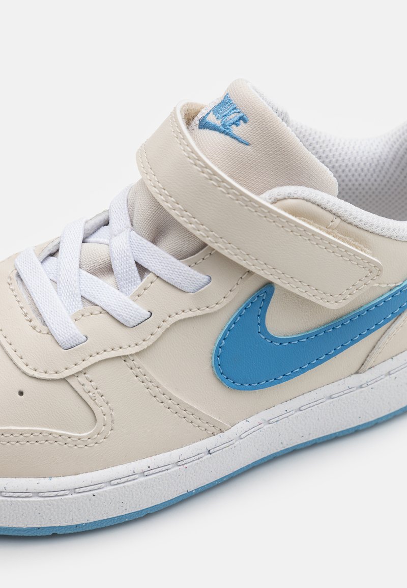 Baskets en cuir beige avec un swoosh Nike bleu, lacets blancs et sangle Velcro. Présentent une surface texturée et une semelle en caoutchouc blanche.