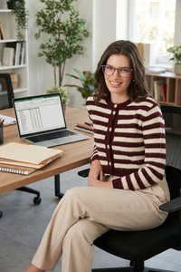 Jeune femme portant des lunettes et un pull rayé, assise sur une chaise de bureau près d’un bureau avec un ordinateur portable affichant un tableur et des dossiers empilés.