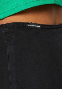 Mustat farkut, joissa on sileä pinta ja näkyvä etiketti, jossa lukee "Marc O'Polo DENIM" vyötäröllä. Vihreä paita näkyy osittain.