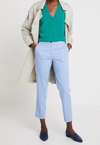 Jasnoniebieskie dopasowane spodnie w połączeniu z zieloną bluzką i beżowym trenczem. Niebieskie teksturowane loafersy dopełniają stylizację.
