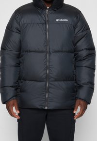 Černá zateplená prošívaná bunda s vysokým límcem, zapínáním na zip a logem Columbia na hrudi. Má prošívaný design a hladký povrch.