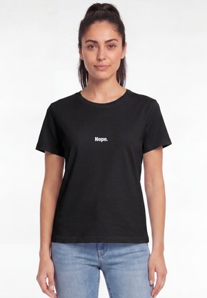 Femme aux cheveux foncés portant un t-shirt noir avec le mot "Nope." en texte blanc, portant un jean bleu clair, debout devant un fond blanc uni.