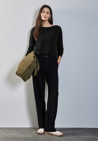 Donna in piedi contro un muro, indossa un maglione nero e pantaloni, tiene una giacca verde oliva, con scarpe da ginnastica bianche e orecchini a cerchio.