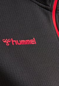 Zwarte textuurstof met een geribbeld patroon, met een opvallend rood "hummel" logo. Ritsdetail in het kraaggebied.