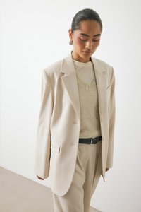 Beige tailored blazer with structured shoulders, paired with a light textured sweater and high-waisted cream trousers, accented by a black belt.