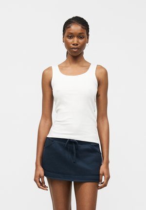 Γυναίκα που στέκεται φορώντας λευκό αμάνικο τοπ και μίνι φούστα μπλε σκούρου με κορδόνι, μπροστά σε απλό φωτεινό φόντο.