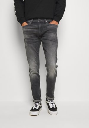 Person trägt ausgewaschene schwarze Slim-Fit-Jeans, an den Knöcheln hochgekrempelt, und schwarz-weiße Sneakers, Hände in den Taschen, vor einfachem Hintergrund.