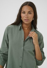 Chemise boutonnee vert clair avec un col, des manches retroussées et une texture subtile. Dotée de boutons bleu clair et d'une coupe décontractée.