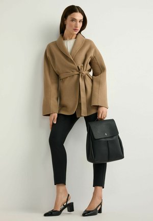 Kobieta ubrana w beżowy płaszcz z paskiem, czarne dopasowane spodnie, czarne sandały na obcasie z paskiem z tyłu, trzymająca czarny skórzany plecak na jasnym, jednolitym tle.