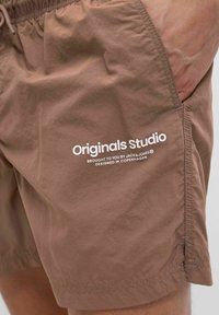 Barna, könnyű anyagból készült sort, elasztikus derékpánttal. Az oldalon fehér nyomtatott szöveg található, amely a "Originals studio" feliratot hordozza.