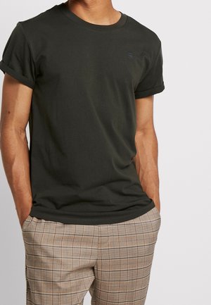 Homme portant un t-shirt noir uni à manches retroussées avec un petit logo et un pantalon beige à carreaux, les mains dans les poches, sur un fond neutre.
