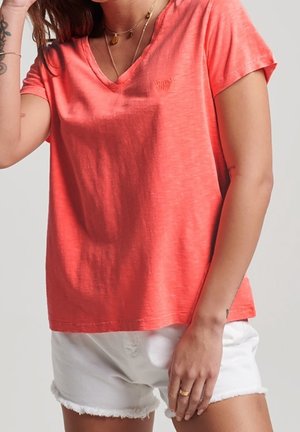 Vrouw die een koraalroze T-shirt met V-hals en korte mouwen draagt, en witte denim shorts met rafelige randen, waarbij een deel van haar kettingen en getatoeëerde armen zichtbaar is.