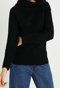 Schwarzer, gestrickter Pullover mit einem breiten, umgeschlagenen Kragen. Besitzt lange Ärmel und eine lockere Passform. Getragen mit blauen Denim-Jeans.