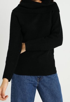 Maglione nero a maglia con scollo largo e ripiegato. Presenta maniche lunghe e una vestibilità rilassata. Indossato con jeans in denim blu.