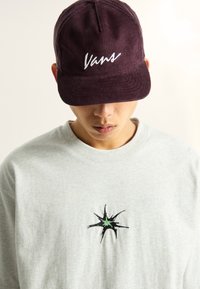 Bordowa czapka z kordurowego materiału z białym haftem "Vans", w zestawieniu z jasnoszarym swetrem z czarnym wzorem gwiazdy z zielonym środkiem.