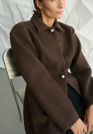 Nő ül egy fémből készült széken, barna bordázott kabátot visel nagy gomb részletekkel, mögötte egy gyűrött fehér háttér.