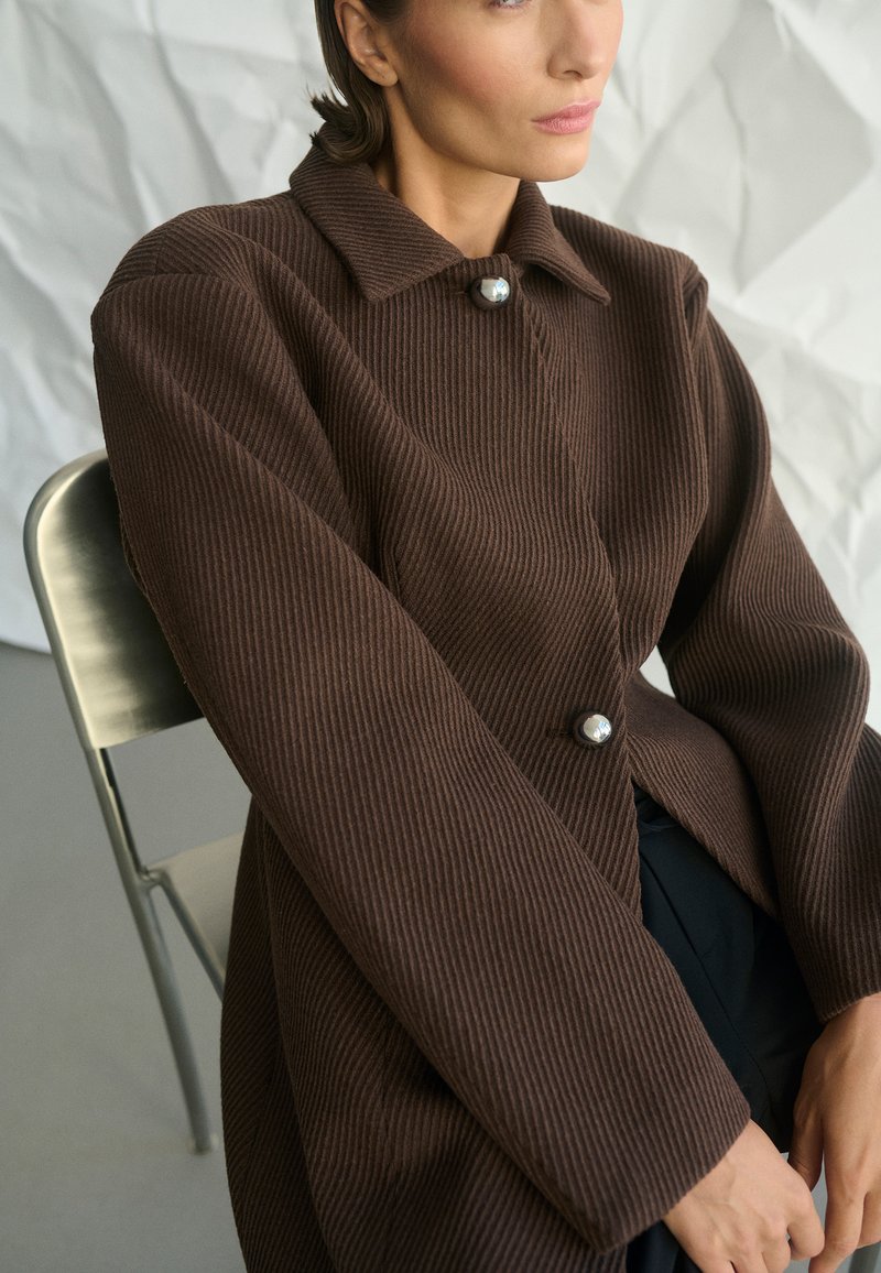 Nő ül egy fémből készült széken, barna bordázott kabátot visel nagy gomb részletekkel, mögötte egy gyűrött fehér háttér.