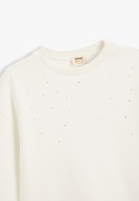 Sudadera blanca con cuello de canalé y acentos plateados dispersos. La tela es suave con una ligera textura.