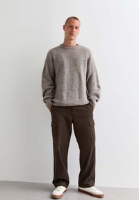 Grijze gebreide trui, oversized pasvorm, ronde halslijn; bruine cargobroek met zijzakken; witte sneakers met tan-zolen; persoon staat met handen in de zakken.