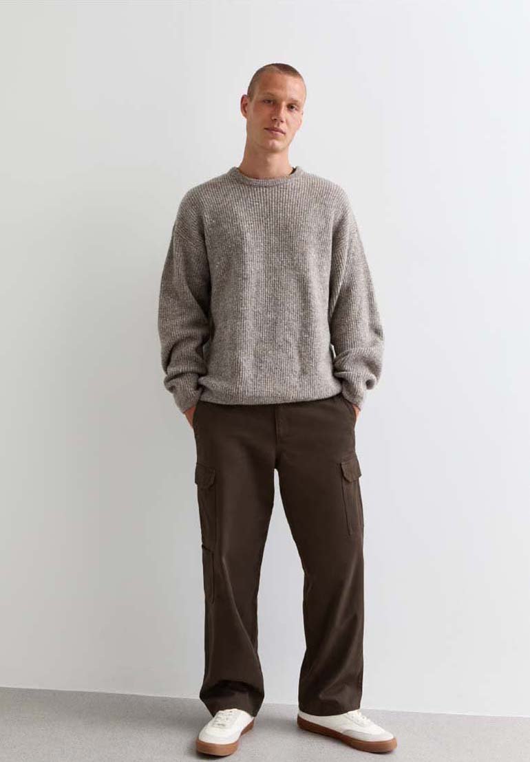 Grijze gebreide trui, oversized pasvorm, ronde halslijn; bruine cargobroek met zijzakken; witte sneakers met tan-zolen; persoon staat met handen in de zakken.