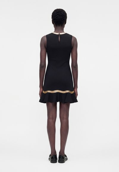 Γυναίκα στέκεται με την πλάτη, φοράει αμάνικο μαύρο φόρεμα με χρυσή διακόσμηση κοντά στο τελείωμα και το λαιμό, συνδυασμένο με μαύρες μοκασίνες.