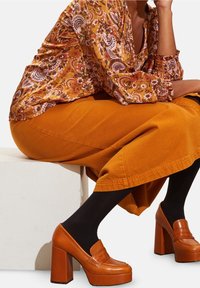 Blusa marrone a fantasia con maniche arricciate, abbinata a una gonna senape. Presenta mocassini arancioni con plateau e collant neri.