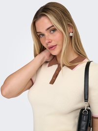 Mlada žena plave kose koja nosi kremasti rebrasti top s smeđim ovratnikom, srebrne naušnice i drži crnu kožnu naramenicu torbe.