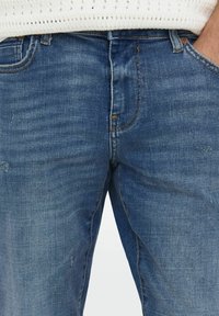 Jeans de mezclilla azul con un diseño clásico de cinco bolsillos, textura desgastada, costuras en contraste y un corte slim. Detalles mínimos de desgaste visibles.