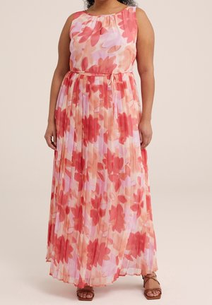 Vrouw draagt een mouwloze, geplooide maxi-jurk met roze en rood bloemenpatroon en een strikceintuur, gecombineerd met bruine sandalen met bandjes.