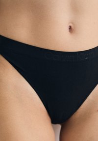 Black Calvin Klein bikini briefs are displayed, highlighting the waistband and a person's midsection, focusing on fabric texture and fit.