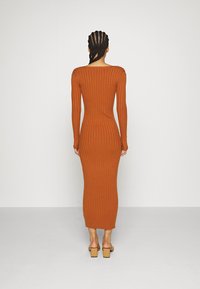 Robe moulante à manches longues côtelée de couleur orange brûlé. Elle présente un décolleté droit dans le dos et une coupe ajustée, portée avec des sandales à talons blocs.