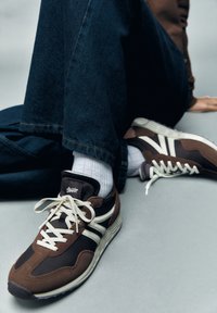 Chaussures de sport marron en daim et mesh, avec des détails et des lacets blancs, posées sur un jean bleu foncé et un fond gris clair.