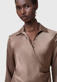 Blusa de satén marrón con cuello, diseño cruzado y cierre de un solo botón. Acabado texturizado que aporta una profundidad sutil. Silueta elegante y ajustada.